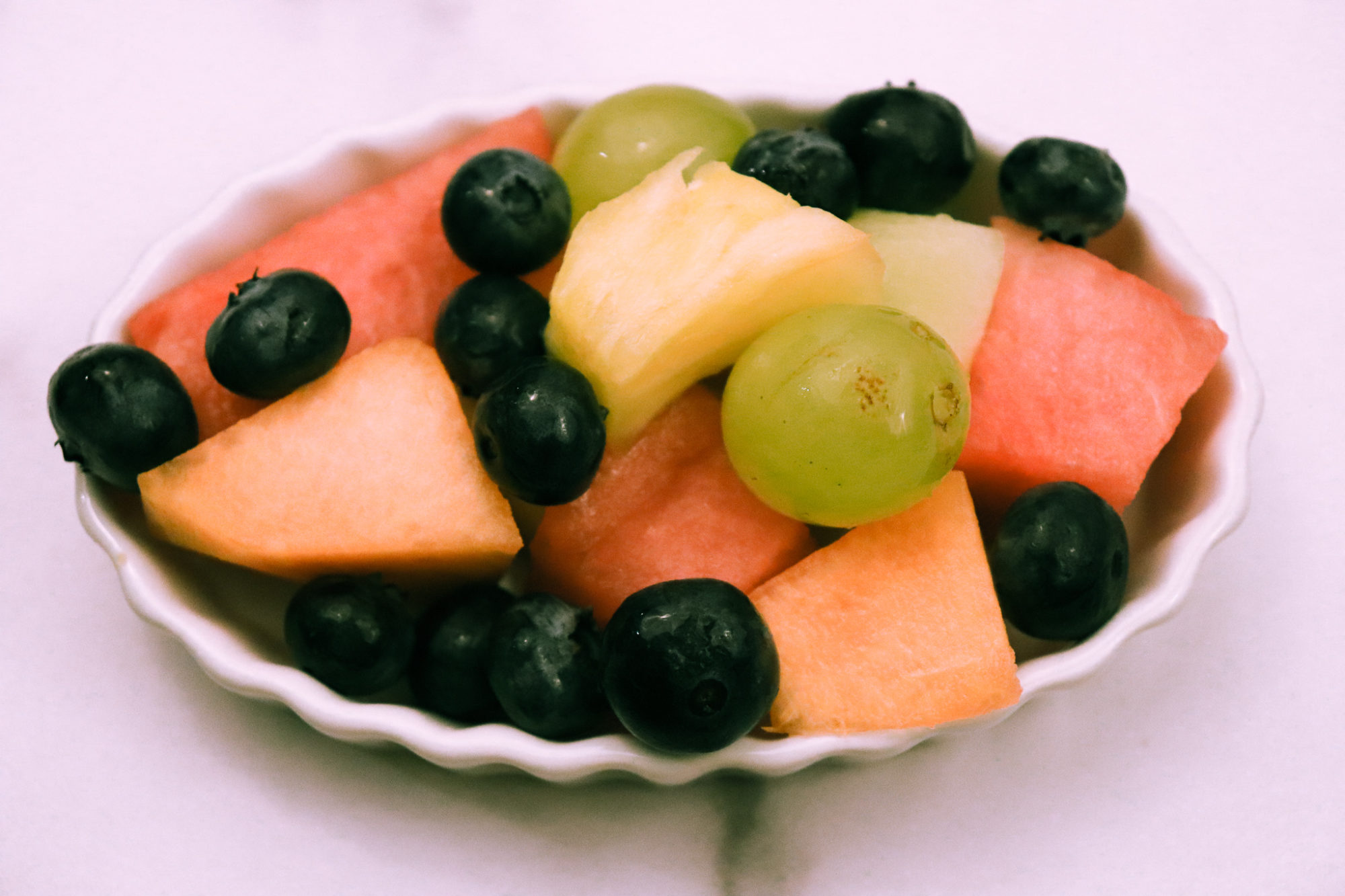 A variety of fresh fruit in a white ramekin on a white marble background.