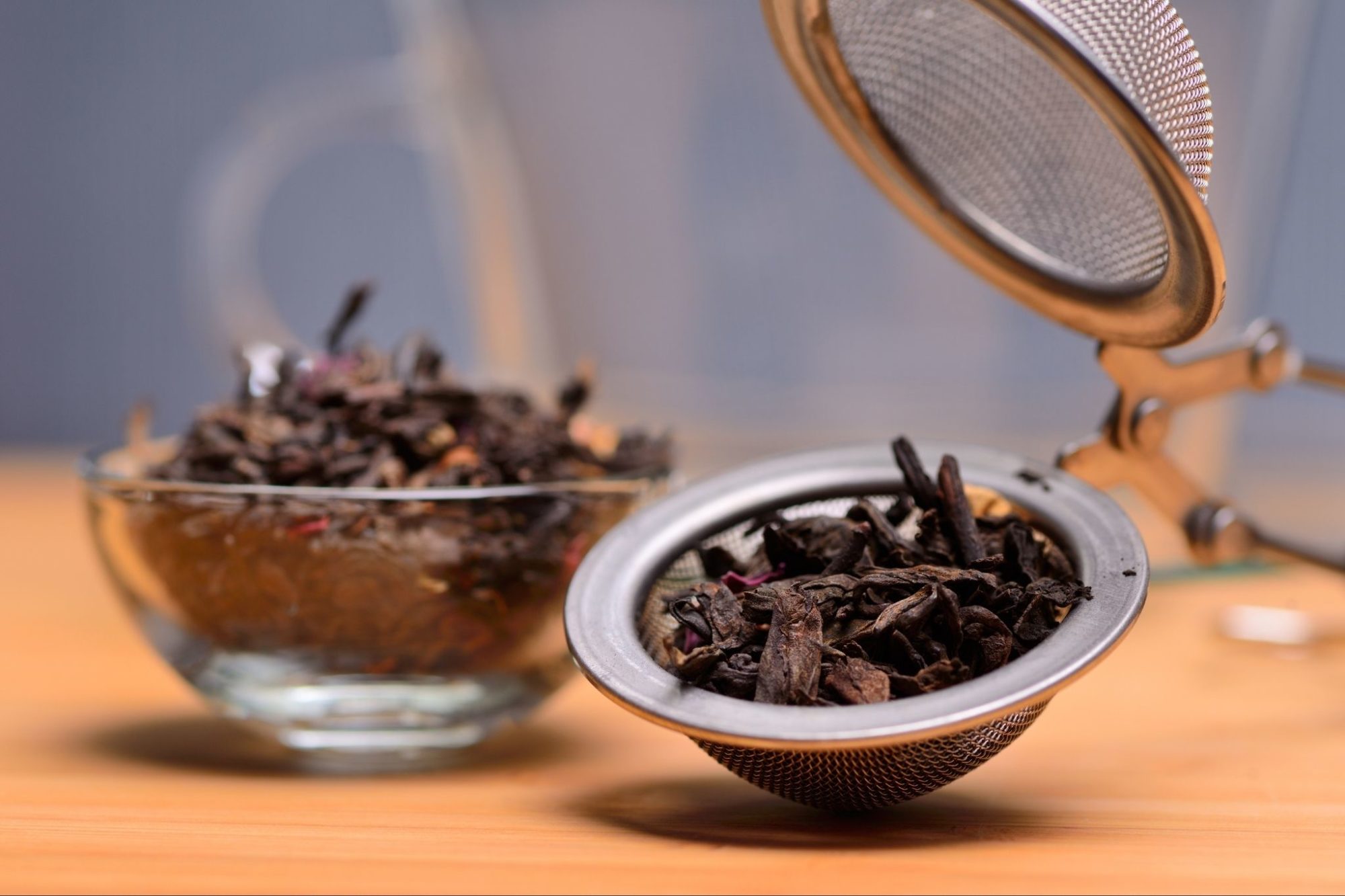Get Started Drinking Tea - Dark tea leaves in a mesh tea ball with a glass bowl of tea leaves in the background