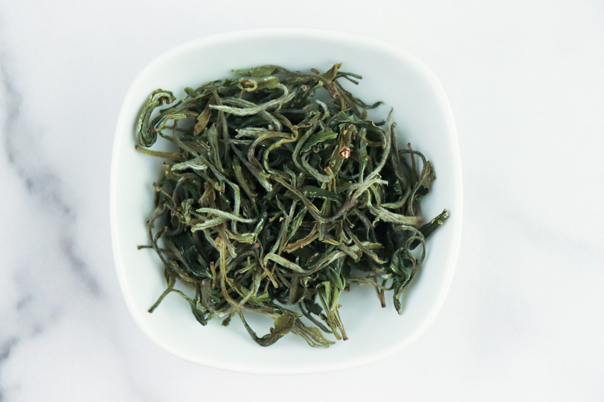 Huang Shan Mao Feng - green tea leaves in a white dish on a white marble background.