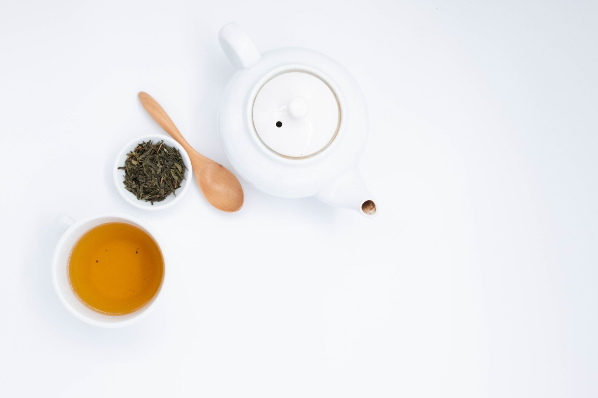 A white tea pot with a bowl of green tea leaves, a wooden spoon, and golden up of tea - Is It Possible to Drink Too Much Tea?